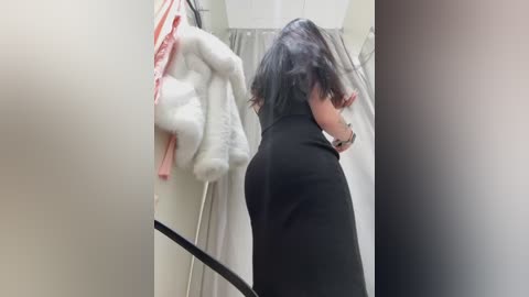 Media: Video of a person with shoulder-length black hair, wearing a black turtleneck, adjusting a white towel in a small, beige-tiled bathroom.