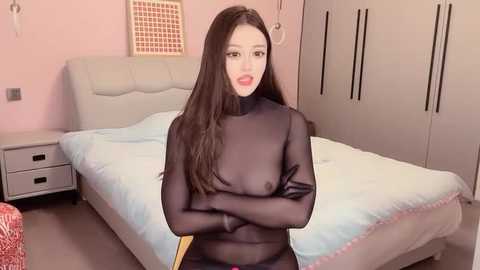 Media: Video of a young Asian woman with long black hair, wearing a sheer black bodysuit, standing in a pastel pink bedroom with a white bed and gray wardrobe.