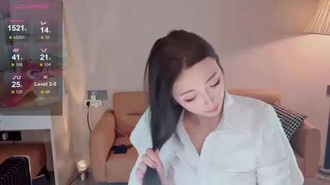 Media: A video of a young Asian woman with long black hair, wearing a white blouse, sitting in a modern living room with beige couches, a TV screen displaying game stats, and a dimly lit room.