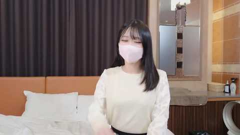 Media: Video of an Asian woman with long black hair, wearing a white mask and long-sleeved shirt, standing near an unmade bed with white sheets, in a modern bedroom with brown and orange walls.