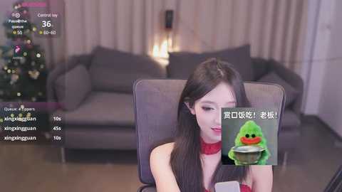 Media: Video of a young East Asian woman with long black hair, fair skin, and a red choker, seated in a modern living room with a gray couch and Christmas lights, wearing a green frog mask.