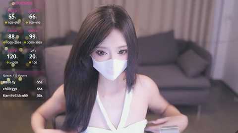 Media: A video of an East Asian woman with long black hair, wearing a white face mask and a white dress, sitting indoors. The background features a grey sofa and beige curtains.