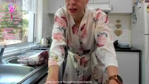 Media: Video of a middle-aged woman with blonde hair, wearing a white floral robe, washing dishes in a modern kitchen with white cabinets, black countertops, and a window displaying greenery.