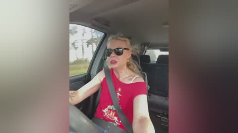 Media: A video of a blonde woman with a slender physique, wearing a red off-shoulder shirt and black sunglasses, driving a car, with a blurred background of trees and a house.