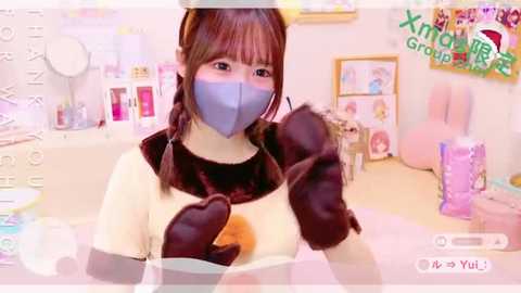 Media: A vibrant, anime-style screenshot from \"Xiao Xiao Group\" shows a young, light-skinned girl with brown hair and a blue mask, wearing a light yellow top and brown gloves, in a playful, pastel-colored room.