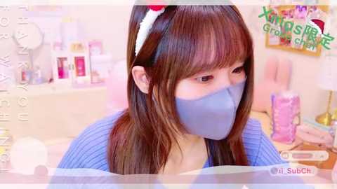 Media: Video of a young Asian woman with long brown hair, wearing a blue mask, seated at a pink desk in a brightly lit room.