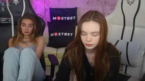 Media: Video of a young woman with long, straight auburn hair, wearing a black top, sitting on a gaming chair in a gaming room with a black \"Mostbet\" banner and purple lighting.