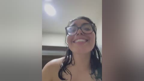 Media: A candid video of a young woman with wet, dark hair, wearing black-rimmed glasses, smiling broadly. She is topless, with a blurred background suggesting an indoor setting.