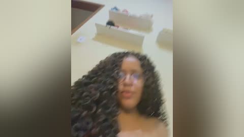 Media: A blurry video shows a young Black woman with curly hair, wearing glasses, standing against a beige wall with a shelf holding toiletries in the background.