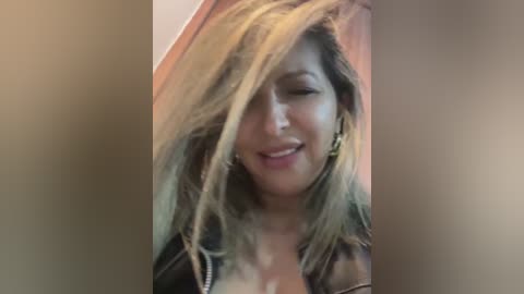 Media: A close-up video of a smiling, light-skinned woman with long, blonde hair, wearing a black leather jacket, captured indoors with a soft, blurred background.