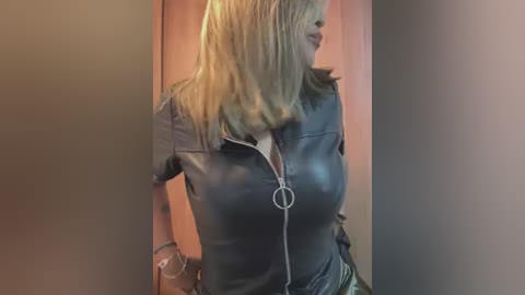 Media: Video of a blonde woman with shoulder-length hair, wearing a tight black leather jacket with a zipper, standing in a dimly lit room with wooden walls.