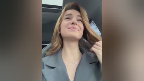 Media: Video of a smiling young woman with light skin and shoulder-length, wavy blonde hair, wearing a light gray blazer. She sits in a car with gray interior, holding a piece of paper.