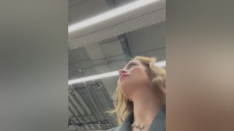 Media: A video of a blonde woman with a fair complexion, wearing a black top, looking upward in an industrial setting with metallic pipes and fluorescent lights.