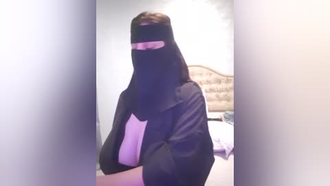 Media: Video of a person in a black niqab with a white t-shirt, seated in a room with a beige tufted headboard and white sheets.