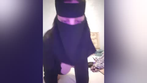 Media: A video of a person wearing a black face mask and a black headscarf, standing in a dimly lit room with a blurred background showing a bed and scattered clothes.