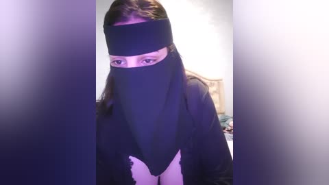 Media: Video of a person with long, dark hair wearing a black headband and face mask, standing in a dimly lit room with blurred, purple-hued furniture and walls.