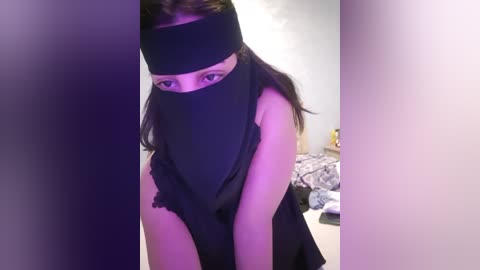 Media: A video of a woman in a black headband, mask, and sleeveless top, bending over in a dimly lit room with a bed in the background.