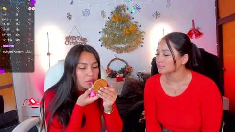 Media: Video of two Asian women with long black hair, wearing red sweaters, eating doughnuts in a cozy, festive room with Christmas decorations and a TV screen displaying a program.