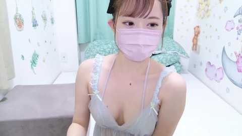 Media: Video of a young East Asian woman with light skin and a slender figure, wearing a light blue lace camisole and pink face mask. She has straight brown hair tied back. Background features pastel-colored decorations and a bed with a green quilt.