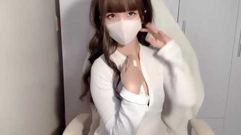 Video of a young East Asian woman with long brown hair, wearing a white face mask, white long-sleeve top, and a bra, sitting in a white chair, indoors.