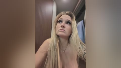 Media: Video of a nude, fair-skinned woman with long blonde hair, looking up thoughtfully. Background shows a wooden door and a dimly lit hallway.