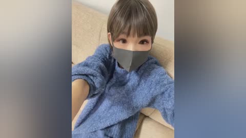 Media: A video of a young Asian woman with short brown hair and fair skin, wearing a blue cardigan and a gray face mask, sitting on a beige couch.