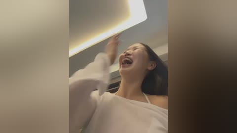 Media: Video of an Asian woman with shoulder-length black hair, wearing a white top, laughing and reaching up, with a modern ceiling light in the background.