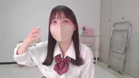 Video of a young Asian woman with long dark hair, wearing a white shirt, red tie, and face mask, in a sterile, white room with a metal ladder and shelves in the background.