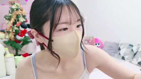 Media: A video of a young East Asian woman with fair skin, black hair in a ponytail, wearing a white mask, grey tank top, and red earrings, sitting indoors with a decorated Christmas tree in the background.