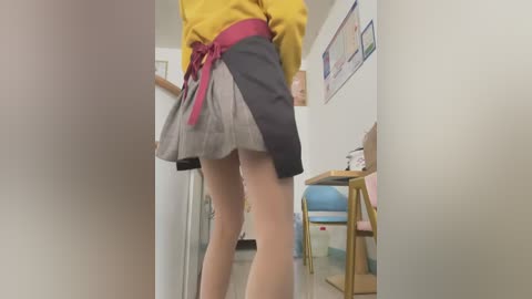 Media: A video of a person wearing a yellow blouse and a short, pleated skirt with a maroon belt, viewed from behind, standing in a cluttered classroom.