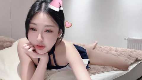 Media: Video of an East Asian woman with long black hair and fair skin, wearing a black bra and skirt, lying on a beige bed with a pink Santa hat, heart emoji, and white walls in the background.