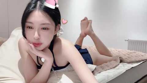 Media: A video of an East Asian woman with long black hair and a slender figure, lying on a bed in a simple room. She wears a black top and shorts, with a pink Santa hat and heart-shaped hair clip.