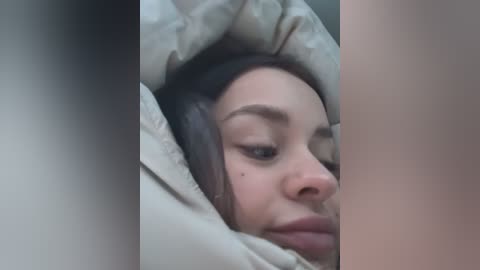 Media: Video of a young woman with light skin, brown hair, and brown eyes, lying in a beige quilt. Her face is partially covered, creating a sense of intimacy and warmth. The background is blurred, emphasizing her expression.