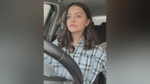 Media: A video of a young woman with long dark hair and fair skin, wearing a light blue and white plaid shirt, driving a car.