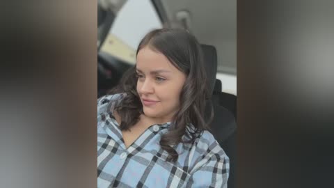 Media: A video of a young woman with long dark hair, wearing a plaid shirt, sitting in a car, smiling and looking slightly to the side.