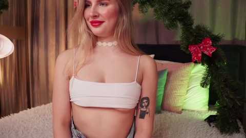 Media: Video of a smiling, fair-skinned woman with blonde hair wearing a white crop top and choker, standing in a cozy, festive room with greenery and pillows.