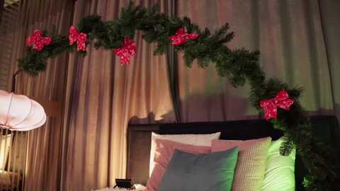 Media: Video of a cozy bedroom with a bed adorned with a green garland, red bows, and pillows in pastel colors. Warm, ambient lighting and beige curtains add to the festive, inviting atmosphere.