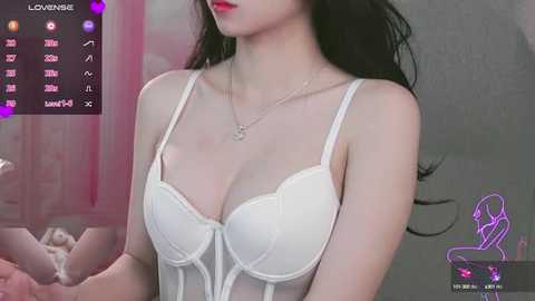 Video of a fair-skinned woman with long black hair, wearing a white lace bra, standing against a blurred background. She has a slender physique and red lipstick.