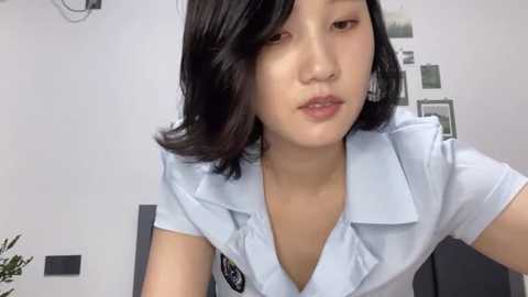 Media: Video of a young East Asian woman with shoulder-length black hair, fair skin, and medium-sized breasts, wearing a light blue blouse with a black badge, sitting indoors.