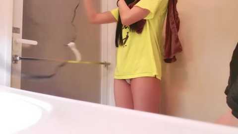 Media: A video of a young woman in a bright yellow t-shirt, with a red towel draped over her shoulder, standing in a bathroom with a white sink and beige walls.