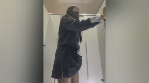 Media: A video of a slim, masked Asian woman with long black hair, dressed in a dark navy school uniform, standing in a narrow, beige-tiled bathroom stall. She holds the door handle, her face partially obscured.