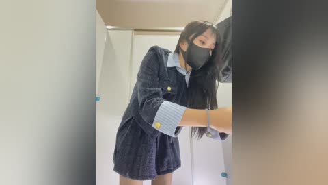 Media: Video of an Asian woman with long black hair and a mask, wearing a blue plaid coat and striped shirt, standing in a public restroom.