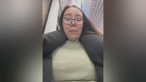 Media: Video of a young woman with glasses, long brown hair, wearing a gray cardigan over a cream sweater, sitting on a subway seat.