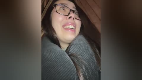 Media: Video of a young woman with glasses, laughing, wrapped in a gray blanket, partially hidden by wooden beams, dimly lit, distressed expression, suggesting confinement.