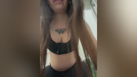 Media: Video of a woman with long brown hair, medium skin tone, wearing a black sports bra, revealing a butterfly tattoo on her chest. Background shows blurred indoor setting.