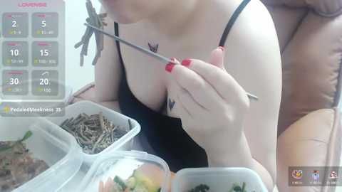 Media: A close-up video shows a fair-skinned woman with a black spaghetti strap top, eating noodles with chopsticks. She has red-painted nails and a small butterfly tattoo on her chest.