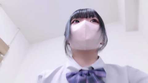 Media: Video of an Asian woman with short black hair, wearing a white shirt with a purple bow tie, a white face mask, and looking upwards. The background is a white, clean room.