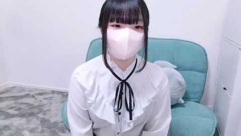 Media: Video of an Asian woman with straight black hair and blunt bangs, wearing a white blouse, black ribbon, and a white mask, sitting on a teal armchair in a minimalist, white room with gray carpet.