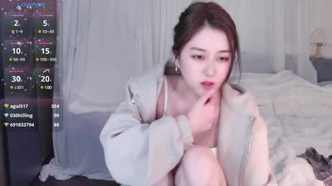 Media: Video of an Asian woman with fair skin, light brown hair, and red lipstick, wearing a beige jacket and a white top, sitting on a bed in a softly lit room.