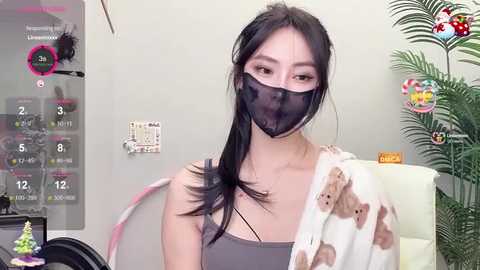 Media: Video of an Asian woman with long black hair, wearing a black face mask, gray tank top, and a white towel with brown bear pattern, in a home setting with a calendar and plants in the background.